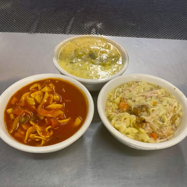 Three bowls of soup: tomato noodle, creamy broccoli, and chicken noodle, on a metal surface.