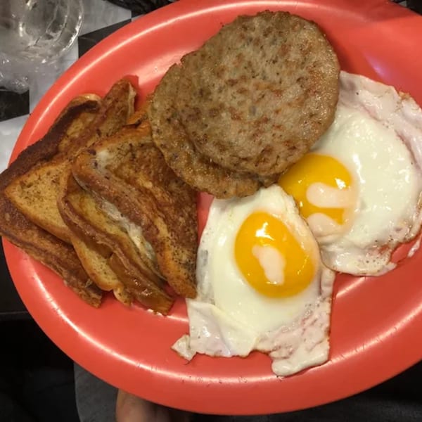 A red plate holds French toast, two fried eggs with runny yolks, and a sausage patty.